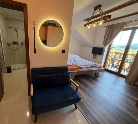Entrance with the bathroom door open to the glass shower, a blue fold-out armchair and the bed by the balcony