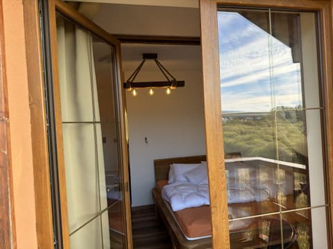The bed and beam lamp seen through the open balcony doors, with the meadow reflected in the glass