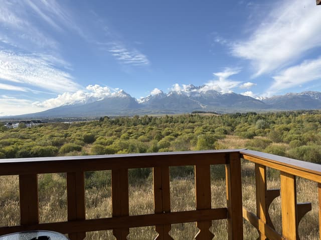View from the apartment balcony – the snow-capped High Tatras and Lomnický štít above a green meadow, framed by the carved wooden railing.